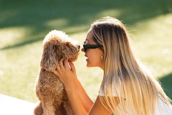A woman in sunglasses nose to nose with a curly-haired dog outdoors
